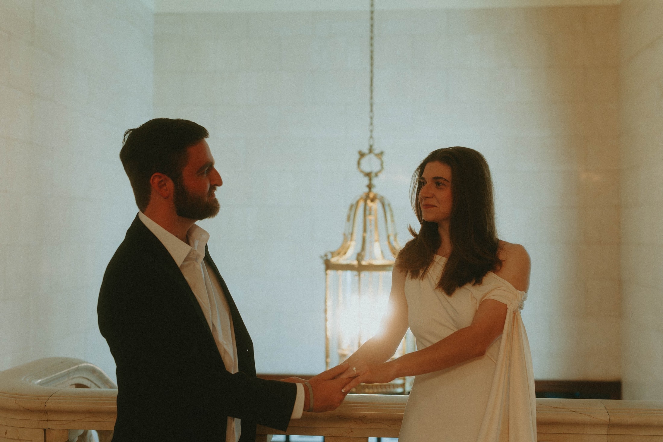 Their hands reaching toward each other beneath a chandelier, rings visible