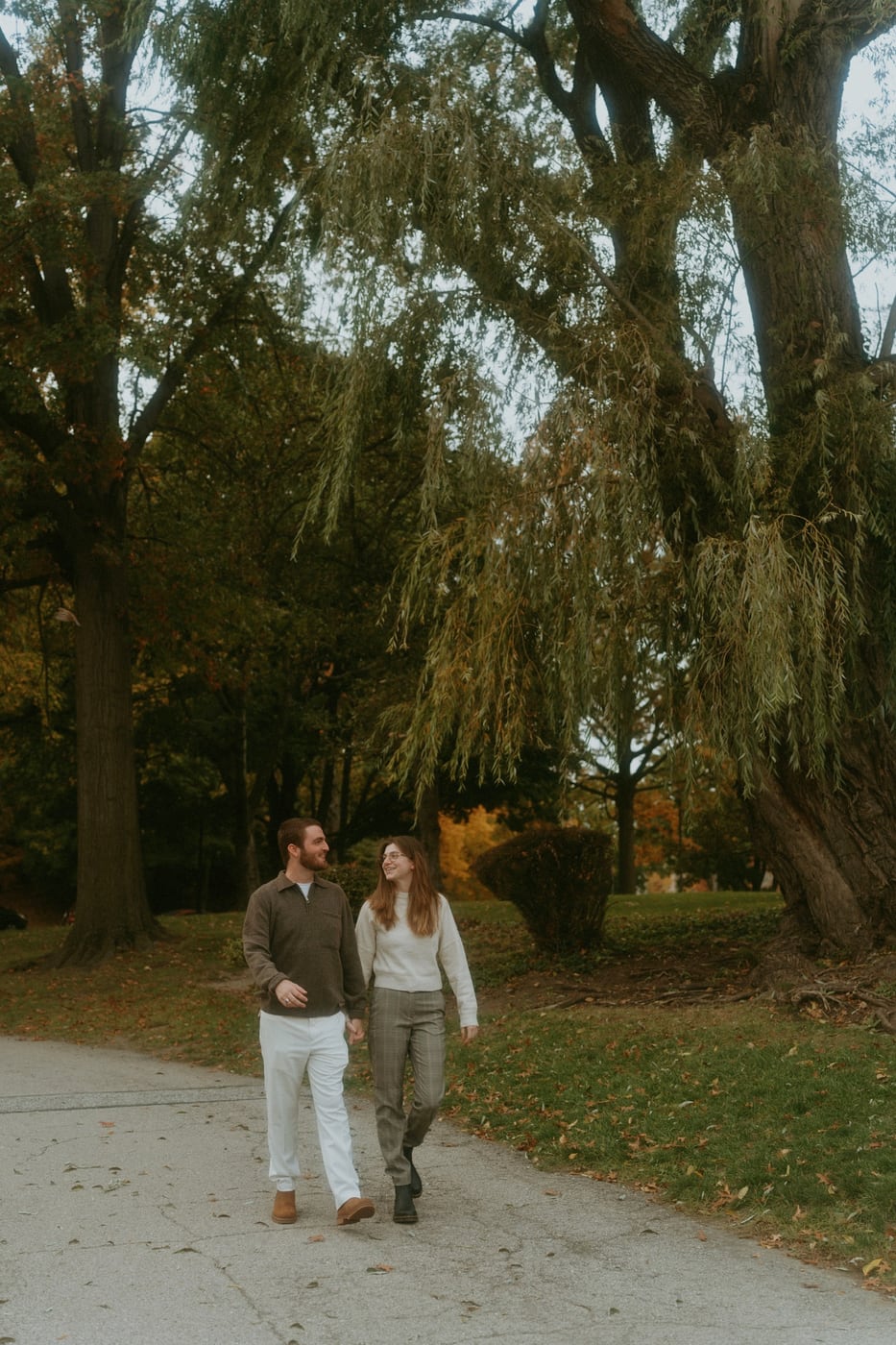 Tylar and Carly walking hand in hand beneath a willow tree in autumn