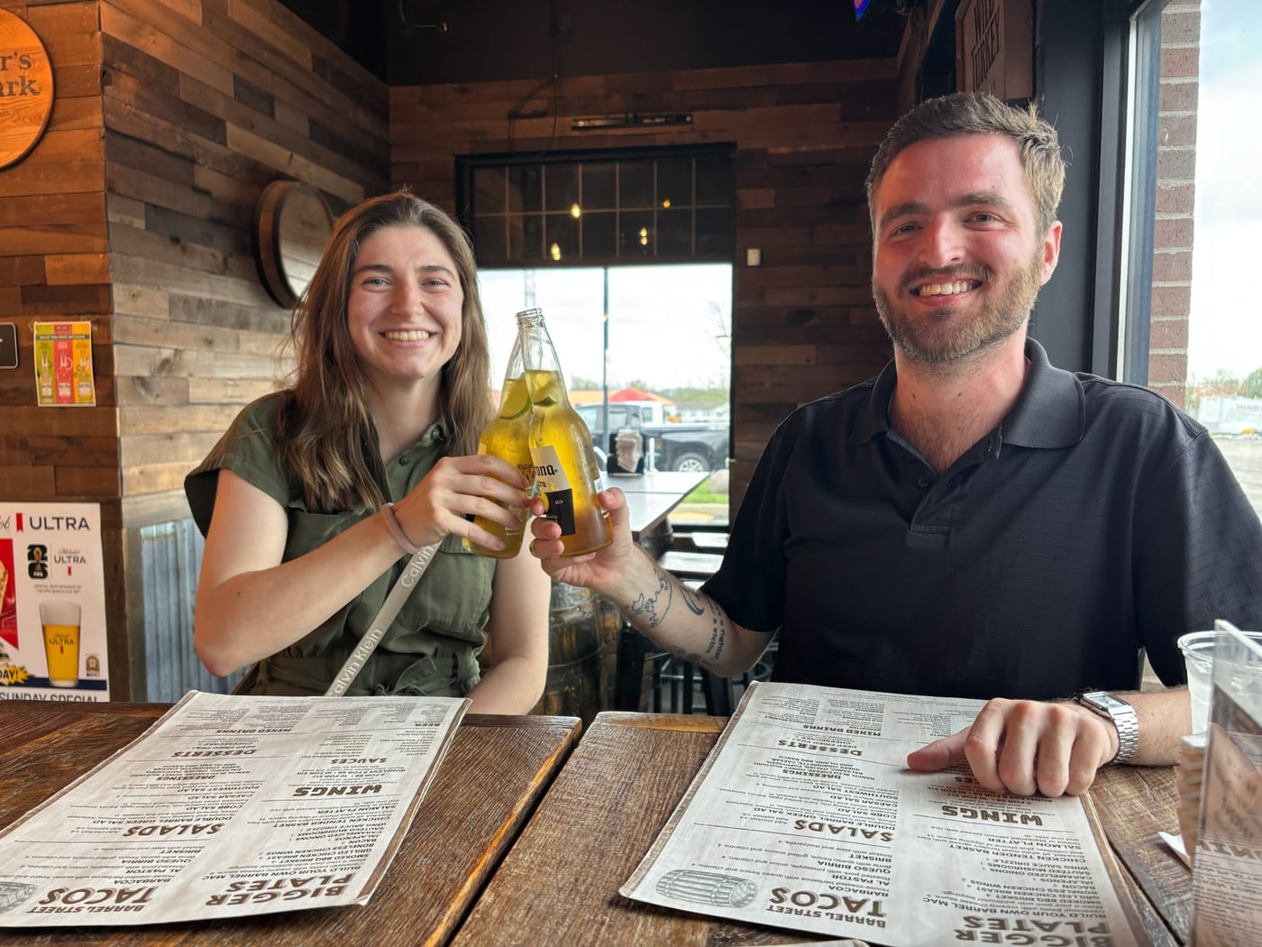 Tylar and Carly clinking bottles across a bar table