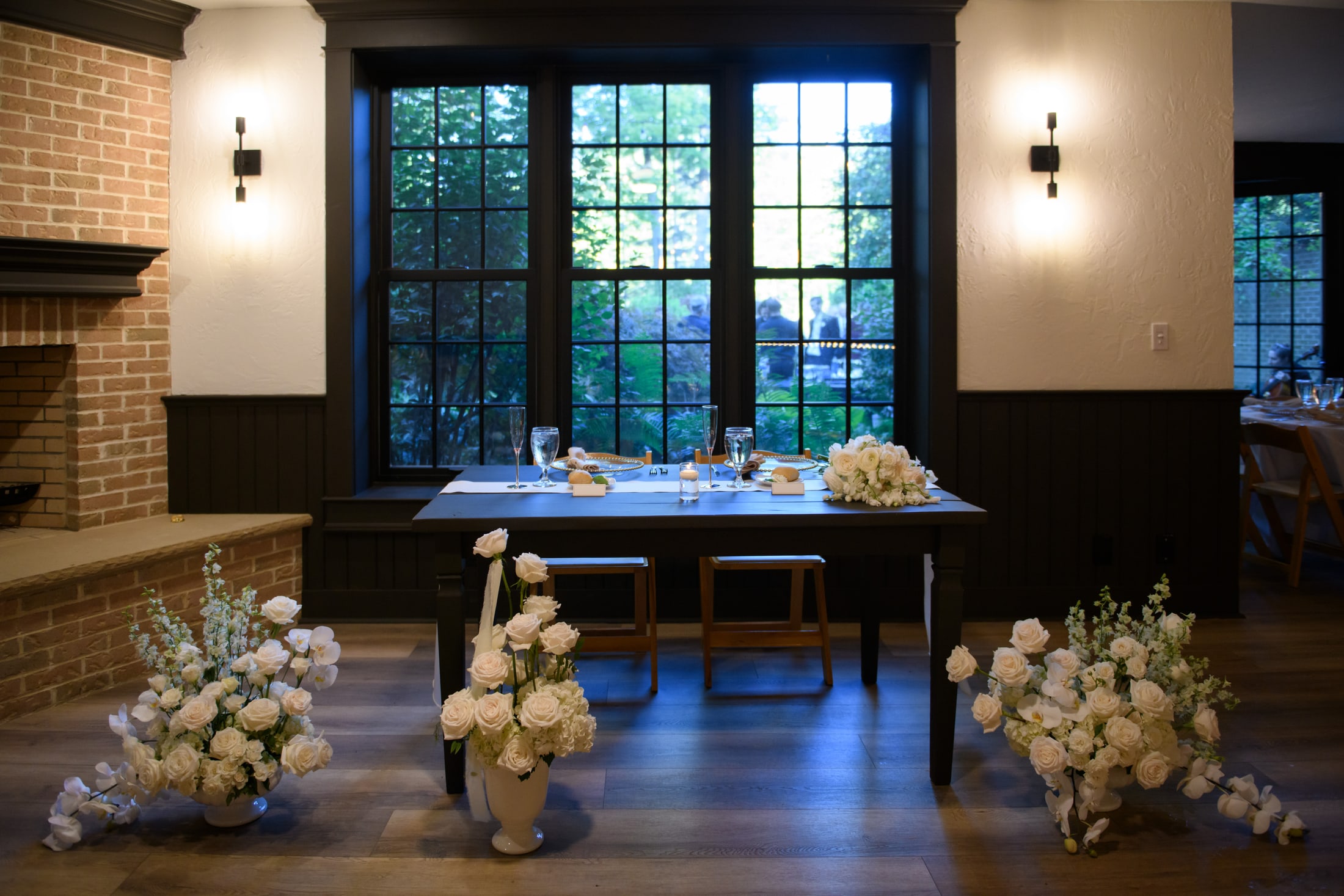 A sweetheart table dressed in white roses before tall dark-framed windows